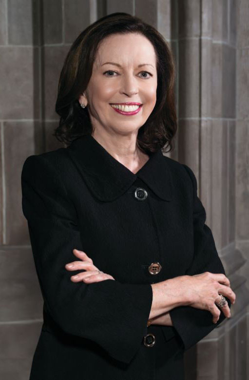 A woman with shoulder-length brown hair, wearing a black buttoned jacket, stands with arms crossed and smiles in front of a stone wall.
