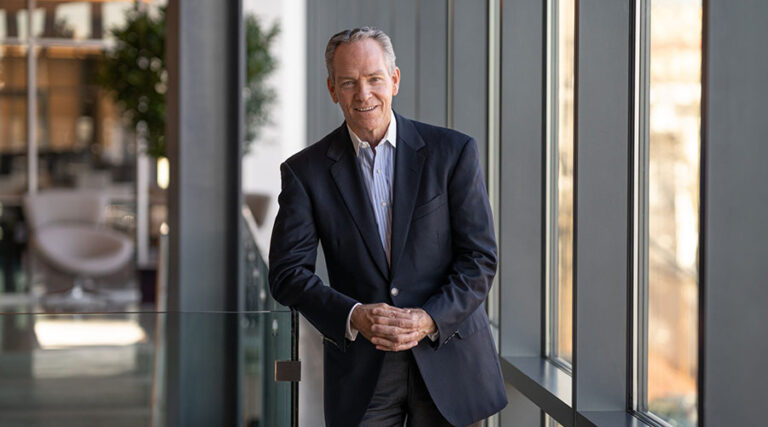 A man in a dark suit stands by large windows in a modern office, smiling and resting his hands on a glass railing. Natural light fills the space, and there are blurred plants and furniture in the background.