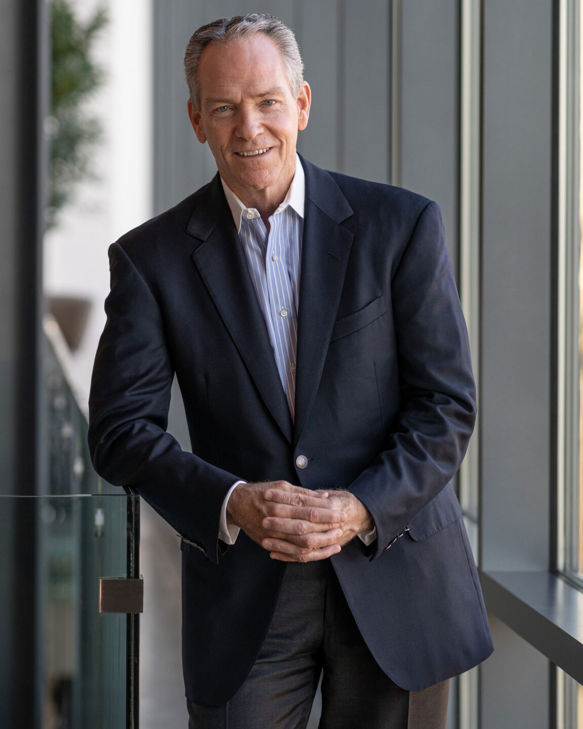A man in a dark suit jacket and striped shirt stands indoors by a large window, smiling and clasping his hands in front of him. The background is softly lit and features glass and greenery.