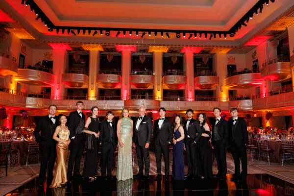 A group of people in formal evening wear pose together on a black floor at an elegant ballroom event with dramatic orange and red lighting and ornate balconies in the background.