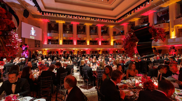 A large, elegant ballroom filled with people in formal attire seated at round tables for a gala dinner, with red and yellow lighting, floral centerpieces, and a stage visible in the background.