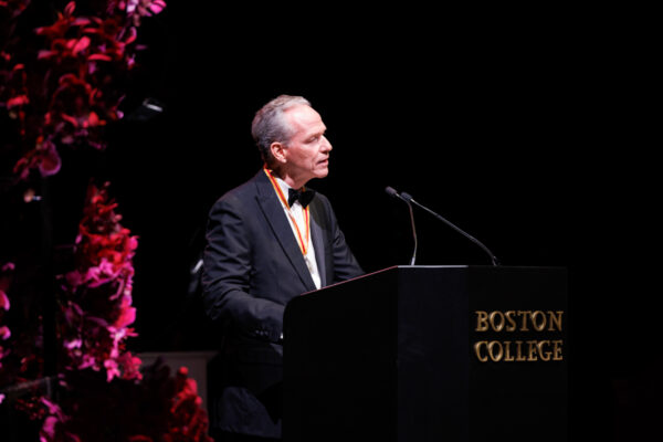 A man in a tuxedo stands at a podium with "Boston College" written on it, speaking into a microphone. Pink flowers are visible to the left, and the background is dark.