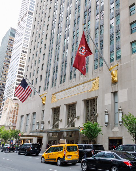 Street view of the Waldorf Astoria hotel in New York City, with American and red flags above the entrance, gold accents, several parked cars, and a yellow taxi in the foreground.