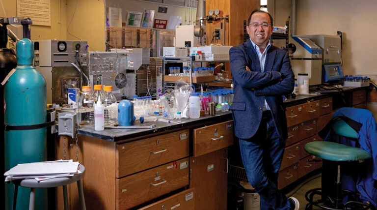 A man in a blazer stands smiling with arms crossed in a laboratory, surrounded by scientific equipment, bottles, and test tubes on cluttered counters.