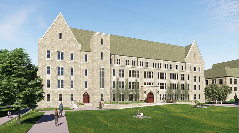 A large stone academic building with tall windows and gabled roofs sits beside a green lawn. People walk on pathways and some sit on the grass under a blue sky.