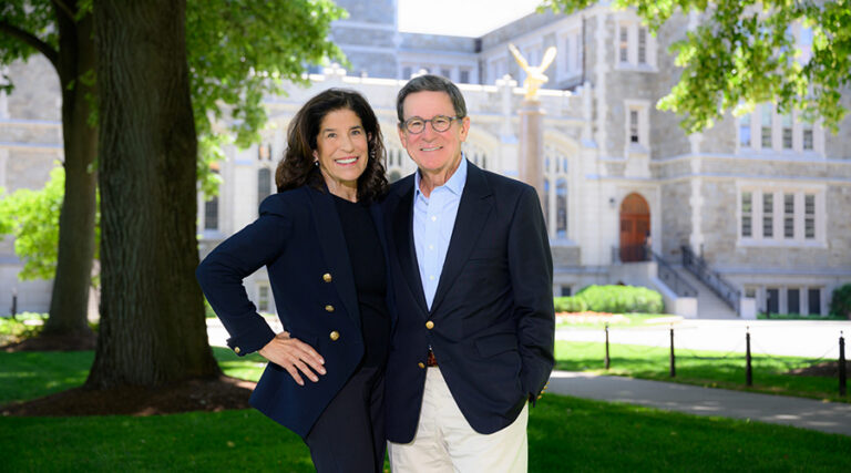 A woman and a man, both in dark blazers, stand smiling together outside on a sunny day, with a large stone building and green trees in the background.