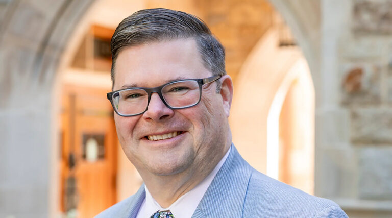 A man wearing glasses and a light-colored suit smiles outdoors in front of a stone building with arches and warm lighting.