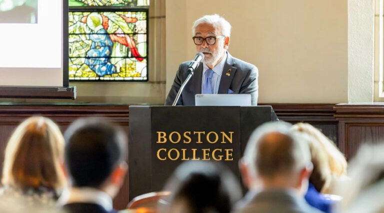 An older man speaks at a podium labeled “Boston College” in a room with stained glass windows, addressing an audience.