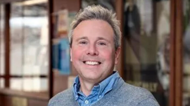 A smiling person with short, light hair wearing a blue collared shirt and gray sweater, standing indoors with blurred posters and windows in the background.