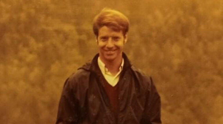 A man with light brown hair smiles at the camera, wearing a dark jacket over a collared shirt and sweater. The background is blurred, featuring muted green foliage under warm, yellow lighting.